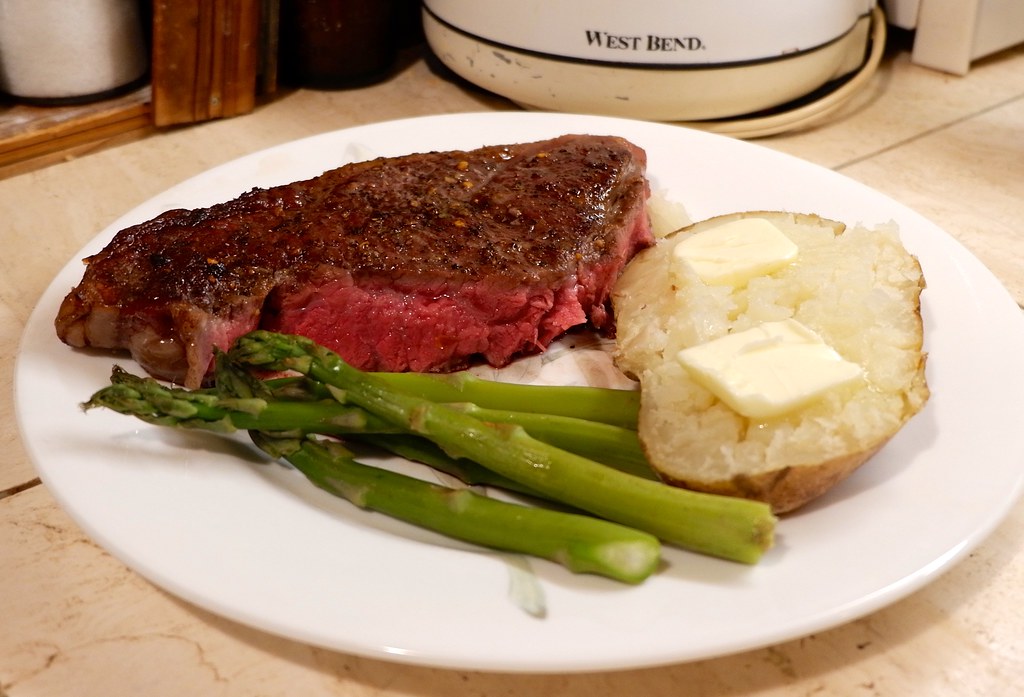 A seared steak resting on a plate with asparagus and a baked potato